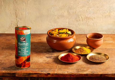 SatRas wood-pressed black mustard oil tin beside a clay jar of mango pickle and brass dishes of chilli, turmeric and fenugreek