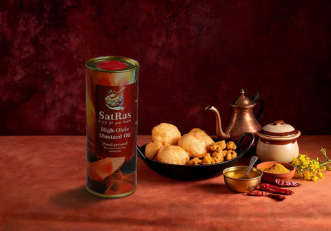 SatRas wood-pressed high oleic mustard oil tin beside a kadai of puris and pakoras and a brass bowl of ghee