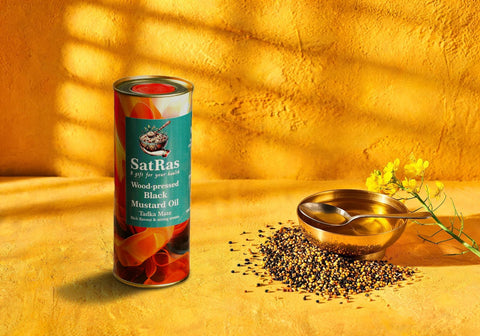 SatRas wood-pressed black mustard oil tin beside a brass bowl of mustard oil, mustard seeds and mustard flowers