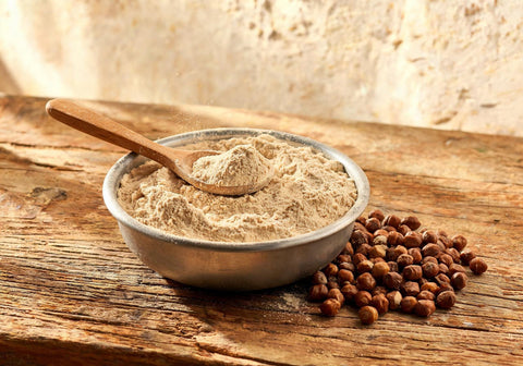 A steel bowl of roasted gram flour, or sattu, with a wooden spoon, beside whole desi black chana on a weathered wooden table