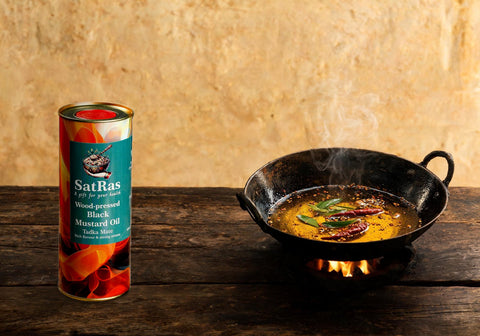 SatRas wood-pressed black mustard oil tin beside an iron kadhai of mustard seeds crackling in hot oil with curry leaves and dried red chillies