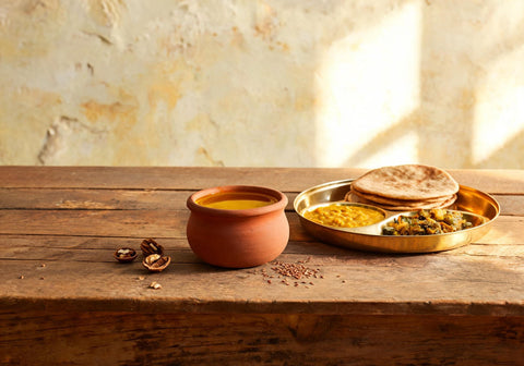 Golden mustard oil in an earthen clay pot beside a brass thali of dal, roti and sabzi with flaxseed and walnuts on a wooden table