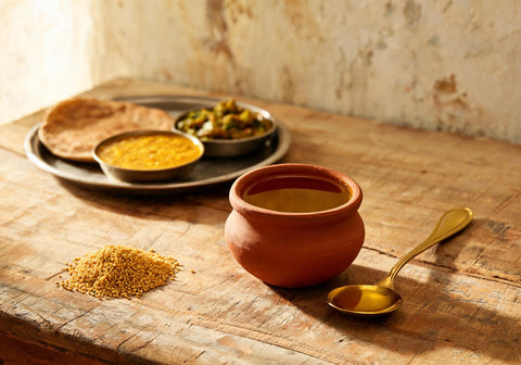 A clay pot of mustard oil and a brass spoon beside a thali of dal, roti and sabzi on a wooden kitchen table