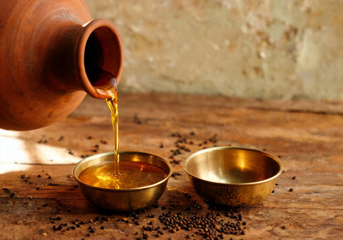Mustard oil poured from a clay pot into a brass measuring katori on a wooden table with scattered mustard seeds