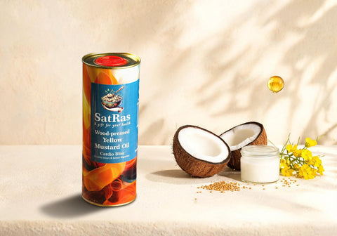 SatRas wood-pressed yellow mustard oil tin on a cream backdrop beside a halved coconut, a jar of coconut oil and yellow mustard flowers