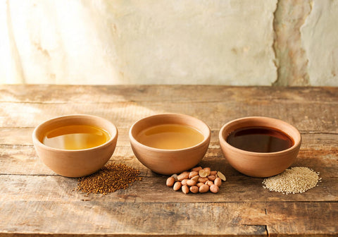 Three clay bowls of cooking oil side by side with mustard seeds, groundnuts and sesame seeds beside each on a wooden table